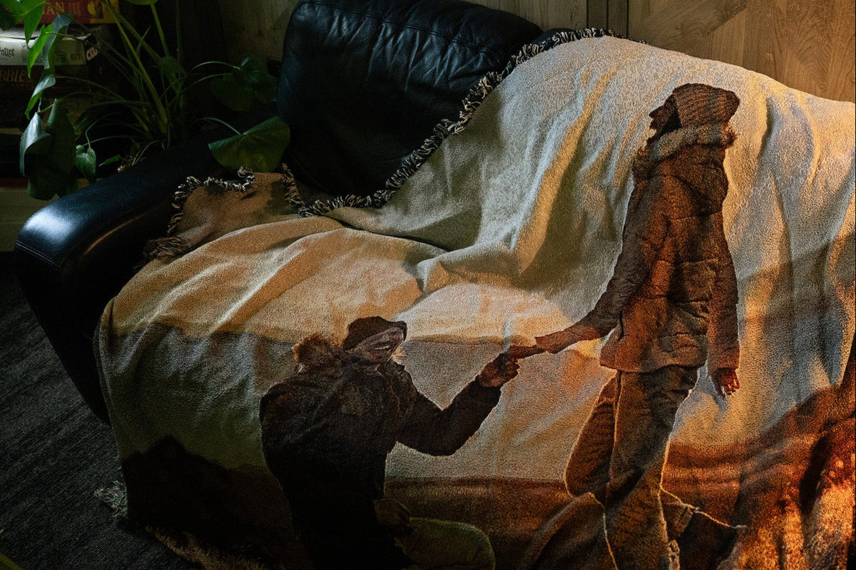 A custom jacquard woven blanket displayed on a black leather couch in a moody, warmly lit living room. The blanket features a woven image of a meaningful moment between two people holding hands, with natural fringe trim and rich warm tones. Board games and a monstera plant visible in the background.