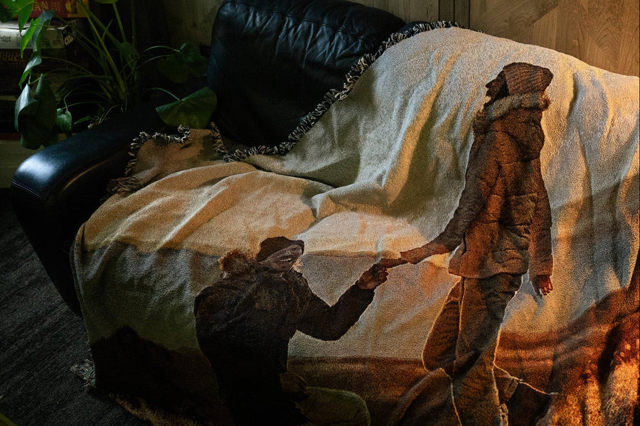 A custom jacquard woven blanket displayed on a black leather couch in a moody, warmly lit living room. The blanket features a woven image of a meaningful moment between two people holding hands, with natural fringe trim and rich warm tones. Board games and a monstera plant visible in the background.